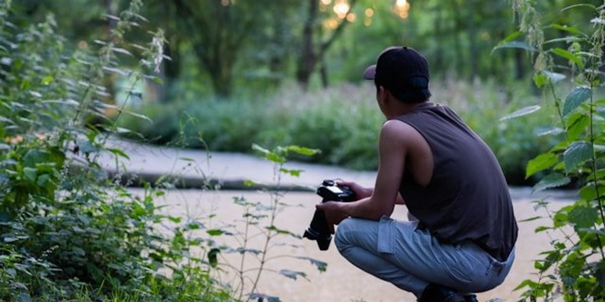 Young South American man crouching at a river with a camera.