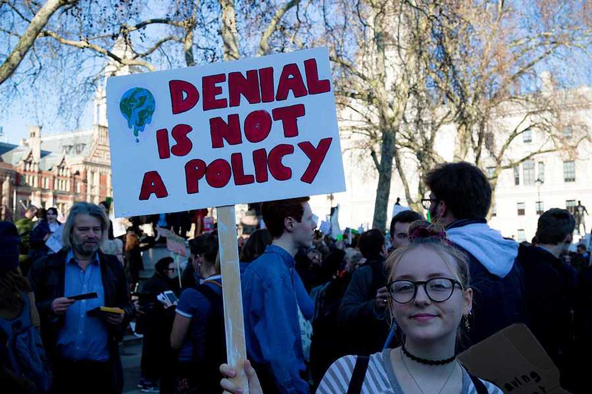 Young woman holding sign of protest reading "DENIAL IS NOT A POLICY."