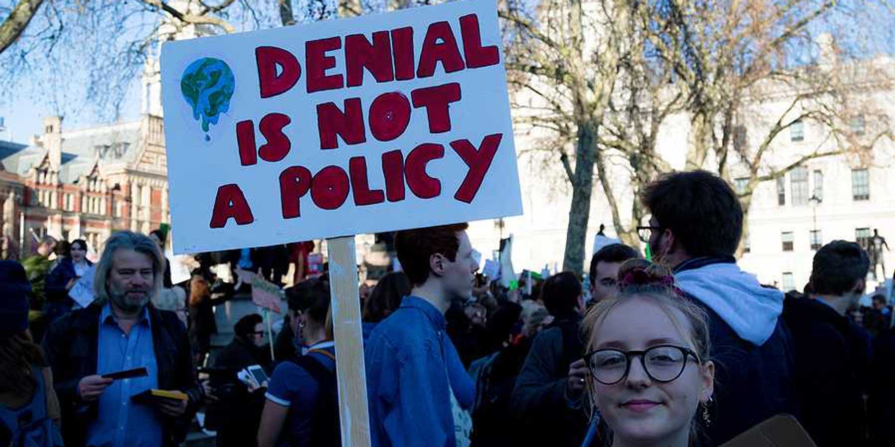 Young woman holding sign of protest reading "DENIAL IS NOT A POLICY."