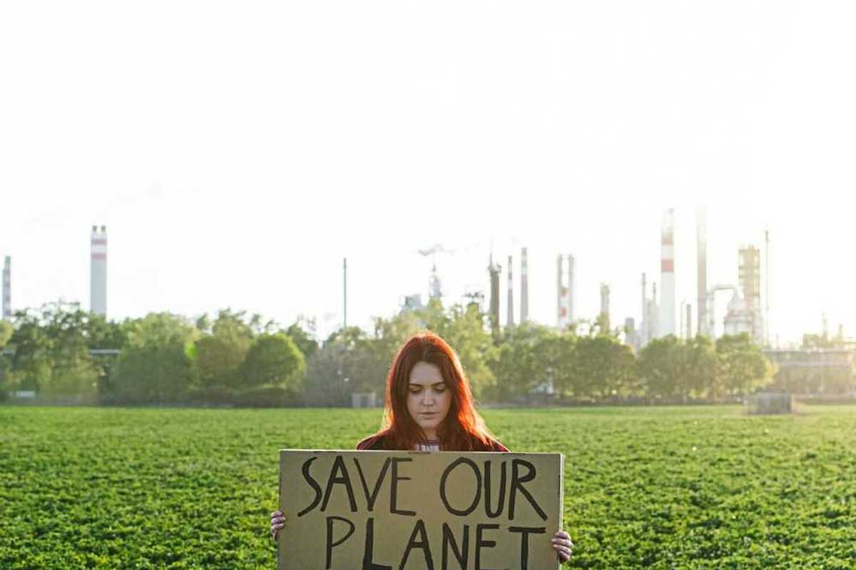 Young woman with polluting power plant in background holds a sign: "SAVE OUR PLANET"