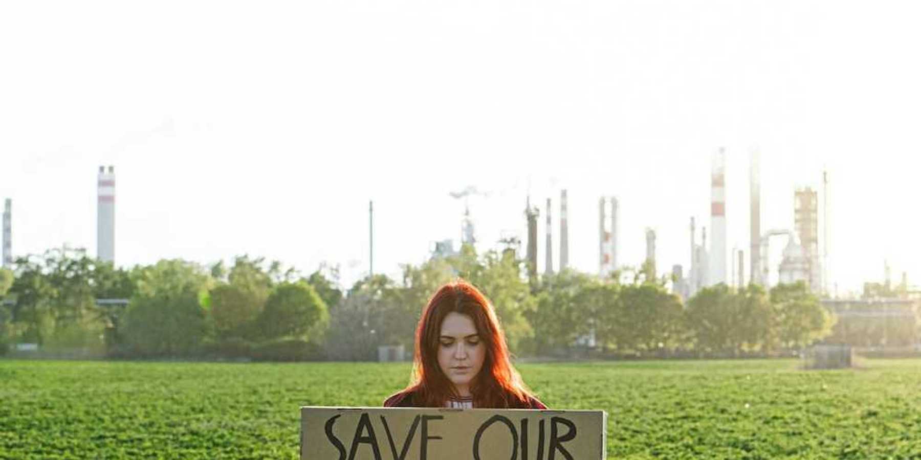 Young woman with polluting power plant in background holds a sign: "SAVE OUR PLANET"